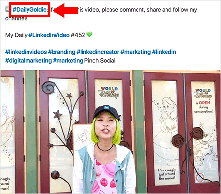 Ini adalah screenshot yang menggambarkan bagaimana Goldie Chan menggunakan hashtag di teks postingan video LinkedIn-nya. Info berwarna merah mengarah ke hashtag #DailyGoldie di teks, yang unik untuk postingan videonya dan membantunya membagikan lagu. Postingan tersebut juga menyertakan hashtag relevan lainnya yang membantu orang-orang menemukan videonya, termasuk #LinkedInVideo. Dalam gambar video, Goldie berdiri di depan beberapa pintu di layar World of Disney. Dia wanita Asia dengan rambut hijau. Dia mengenakan topi LinkedIn hitam, kalung choker hitam, kemeja bermotif macaron merah muda, dan jaket biru dan putih.
